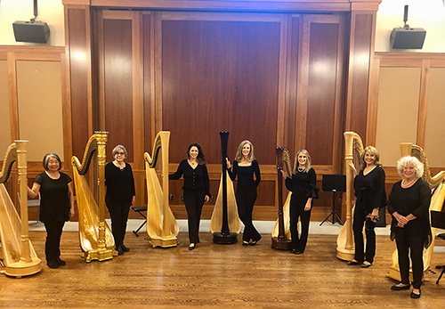 The Harpworks Ensemble - 7 Harps and 7 Harpists standing in a semi- circle in a wood-paneled venue