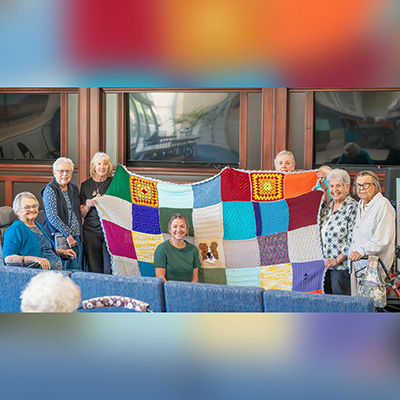 Members of our knitting group shown holding up the handmade quilt presented to Sarah, seated in front, the wife of our Chief Engineer Steve.